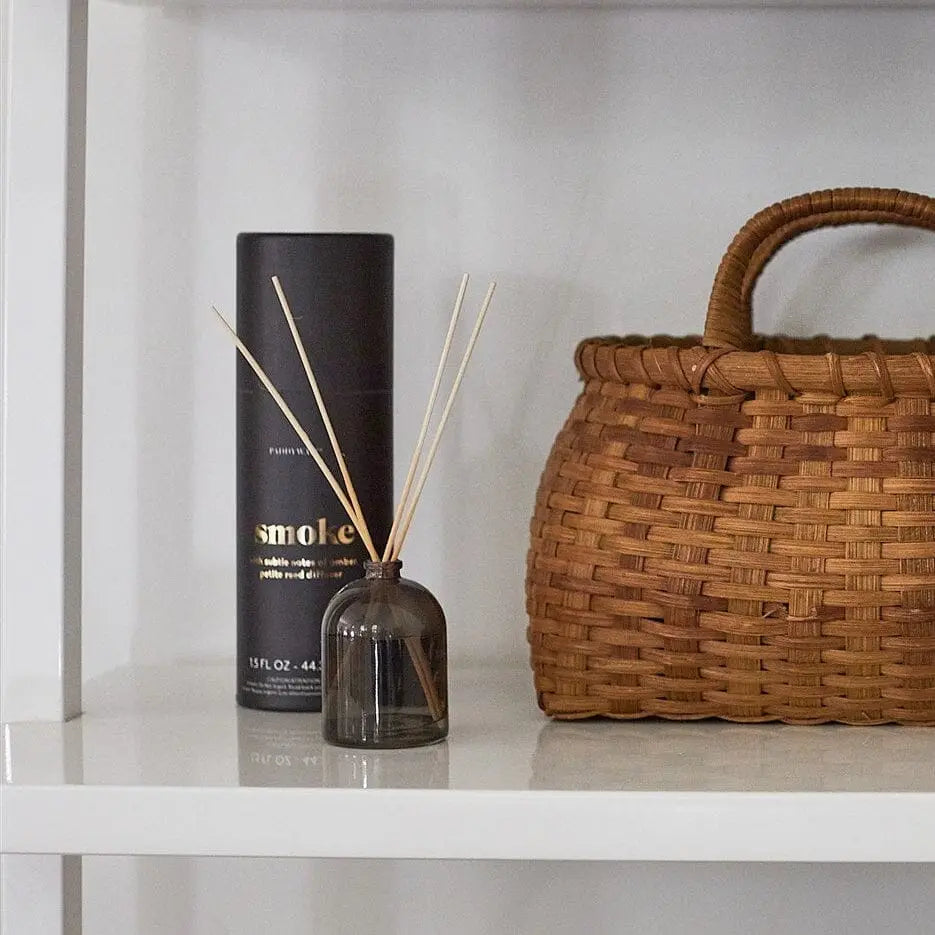 The Smoke - 1.5 oz Diffuser with reed sticks, a black cylindrical container, and a woven handled basket are arranged on a white shelf.