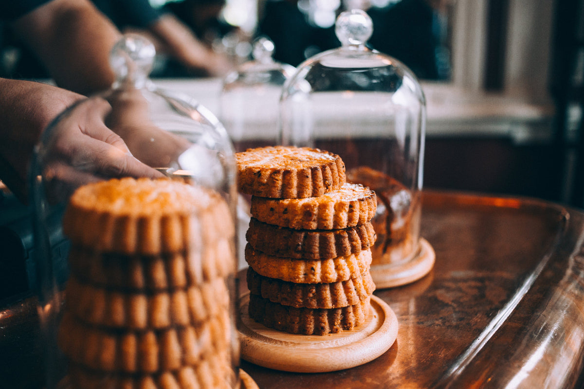 Stacks of round cookies are displayed on wooden trays under glass domes, with a person’s hand reaching toward the cookies. The scene is set indoors on a polished wooden surface.