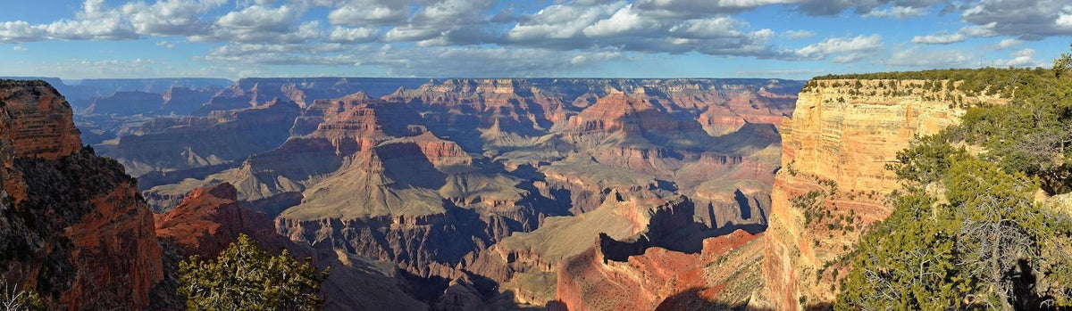 Panoramic view of the Grand Canyon with dramatic rock formations, layered cliffs, and deep valleys under a partly cloudy sky; sunlight highlights the red and orange hues of the landscape.