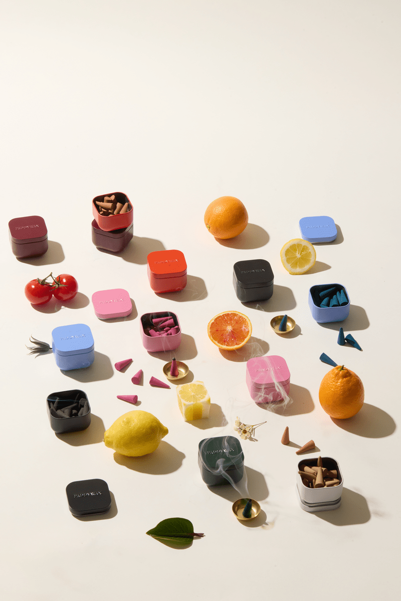 Colorful Pomelo Bay - Metal Incense Tins, filled with triangular cones, are arranged with citrus fruits, tomatoes, leaves, and small scoops on a white surface. The vibrant tins—blue, red, pink, black, and white—evoke the playful packaging often seen in best-selling fragrances or specialty scented incense.