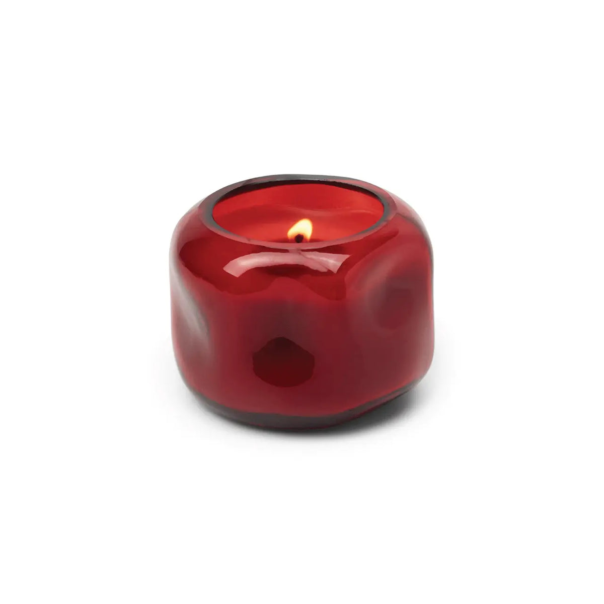 A small, round red glass vessel containing the Yuzu Woods - Basalt 5 oz Candle sits lit against a white background.