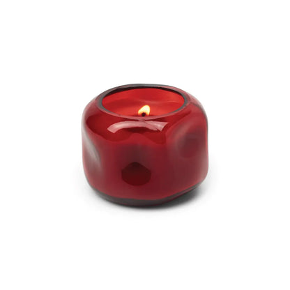 A small, round red glass vessel containing the Yuzu Woods - Basalt 5 oz Candle sits lit against a white background.