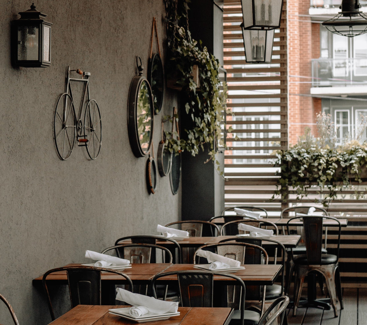A cozy outdoor café with wooden tables, metal chairs, and neatly folded white napkins. The wall is decorated with a mirror, plants, and a metal bicycle. Light streams in through slatted panels, creating a warm ambiance.