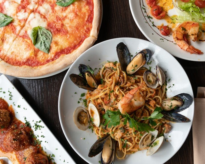 A table with plates of Italian food, including a cheese pizza with basil, pasta with seafood and herbs, meatballs in sauce, and a salad with shrimp, lettuce, and tomatoes.