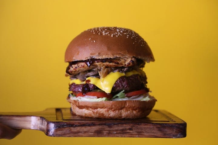 A large cheeseburger with lettuce, tomato, cheese, grilled onions, and meat patties in a sesame seed bun, placed on a wooden board against a yellow background.