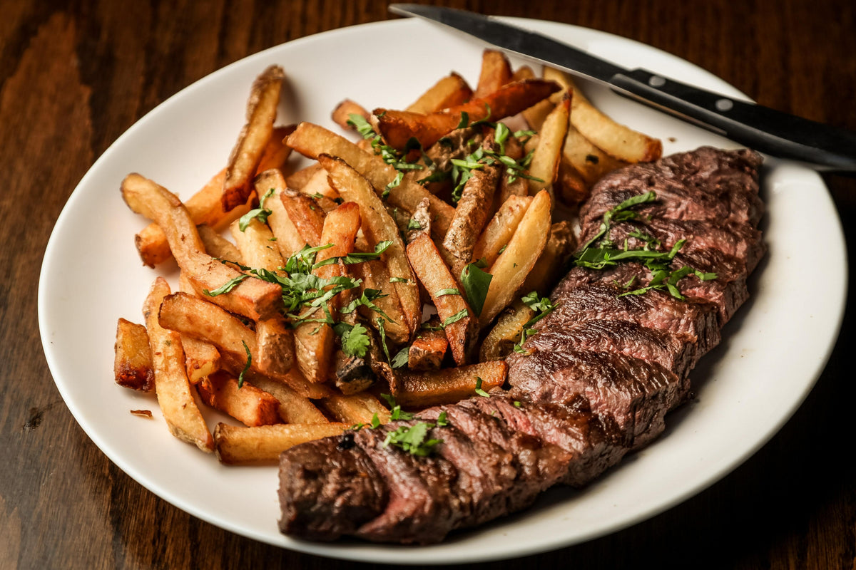 A white plate with grilled steak garnished with herbs and a serving of French fries, also topped with herbs, sitting on a wooden table. A steak knife rests on the edge of the plate.