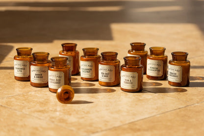 A Sea Salt + Sage - Apothecary 8 oz Candle in an amber glass jar rests on its side in the sunlight, alongside other vintage-inspired fragrance apothecary bottles displayed on a tiled surface.