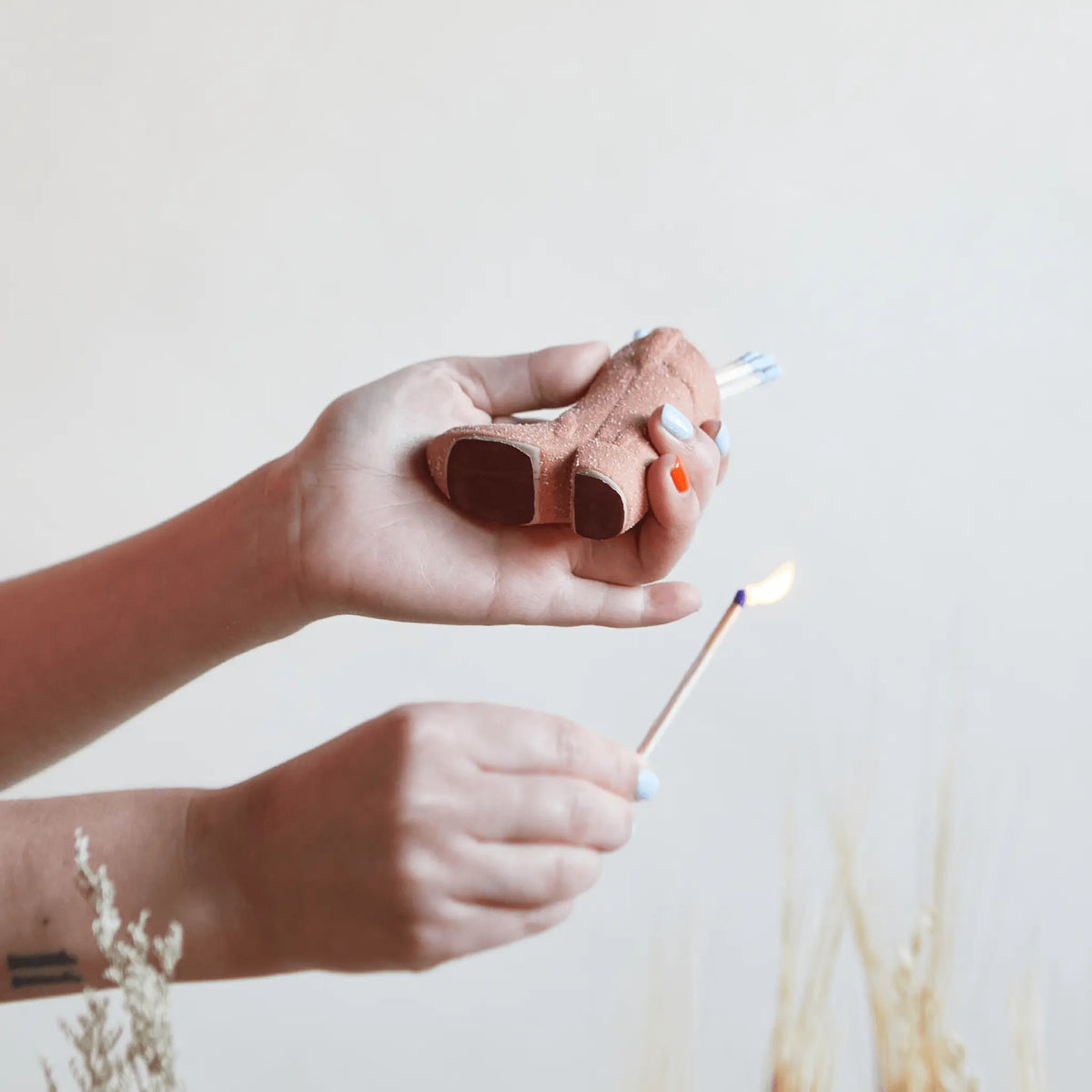 A person holds a white Cowboy Boot Match Holder in one hand and lights a match with the other, set against a neutral background with dried plants—an arrangement that brings subtle Western home decor charm to any space.