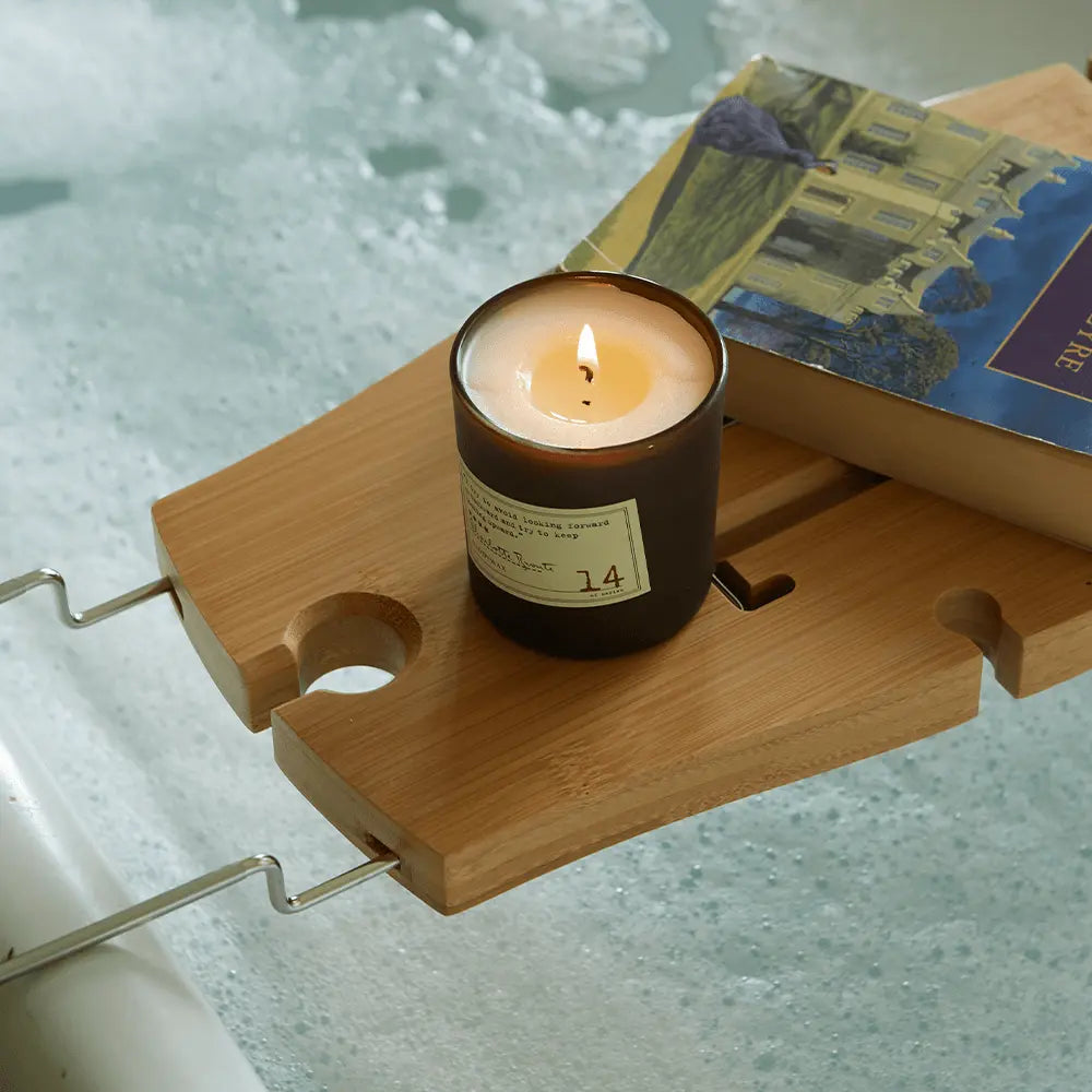 A Charlotte Bronte - Library Candle 6 oz and a book sit on a wooden bath tray above bubbly water, creating the ideal gift experience for fans of the Library Collection fragrance.