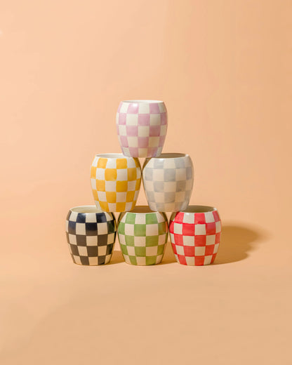 Five Checkmate 11 oz Candles in Cactus Flower are stacked in a pyramid against a peach background. These hand-painted vessels display black, green, red, yellow, pink, and blue checkered patterns, adding a playful touch to your reusable decor.