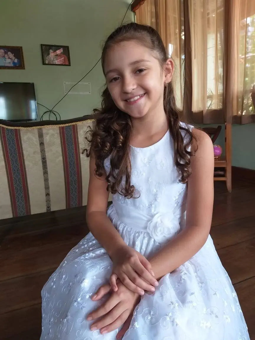 A young girl with long curly hair in a white dress sits smiling indoors on a wooden floor, with a Sky + Rain St. Jude Giveback "Hope" 11 oz. candle and framed photos adding warmth to the background beside a couch and TV.