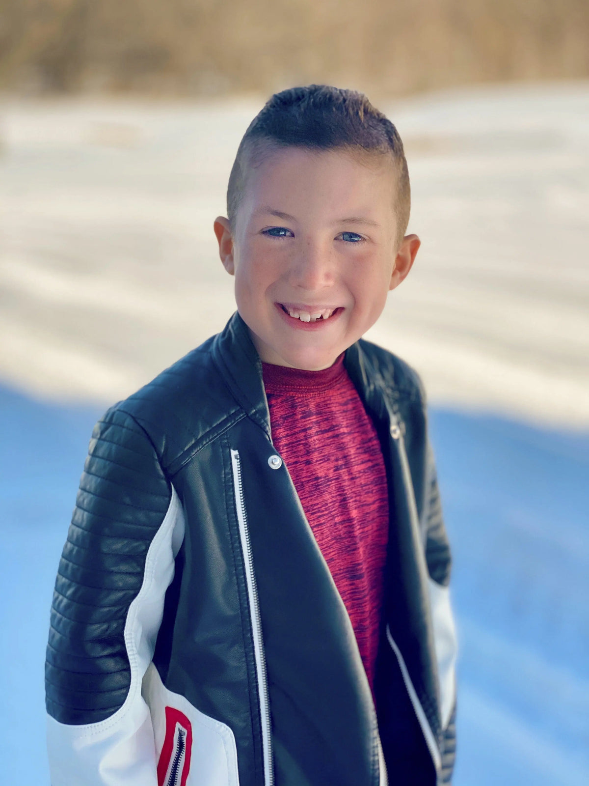 A smiling boy with short brown hair stands outdoors on a snowy day, wearing a black and white jacket over a red shirt, holding the Cactus Flower - St. Jude Giveback "Possibilities" 11 oz. Candle to support childhood cancer awareness.