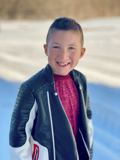 A smiling boy with short brown hair stands outdoors on a snowy day, wearing a black and white jacket over a red shirt, holding the Cactus Flower - St. Jude Giveback "Possibilities" 11 oz. Candle to support childhood cancer awareness.