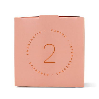 A pink box featuring the number 2 at its center, encircled by the words “CARING,” “INTERPERSONAL,” “GENEROUS,” and “EMPATHETIC” in a circular design—perfect for gifting as the Enneagram #2 Helper 5 oz Candle in Violet + Vanilla fragrance.