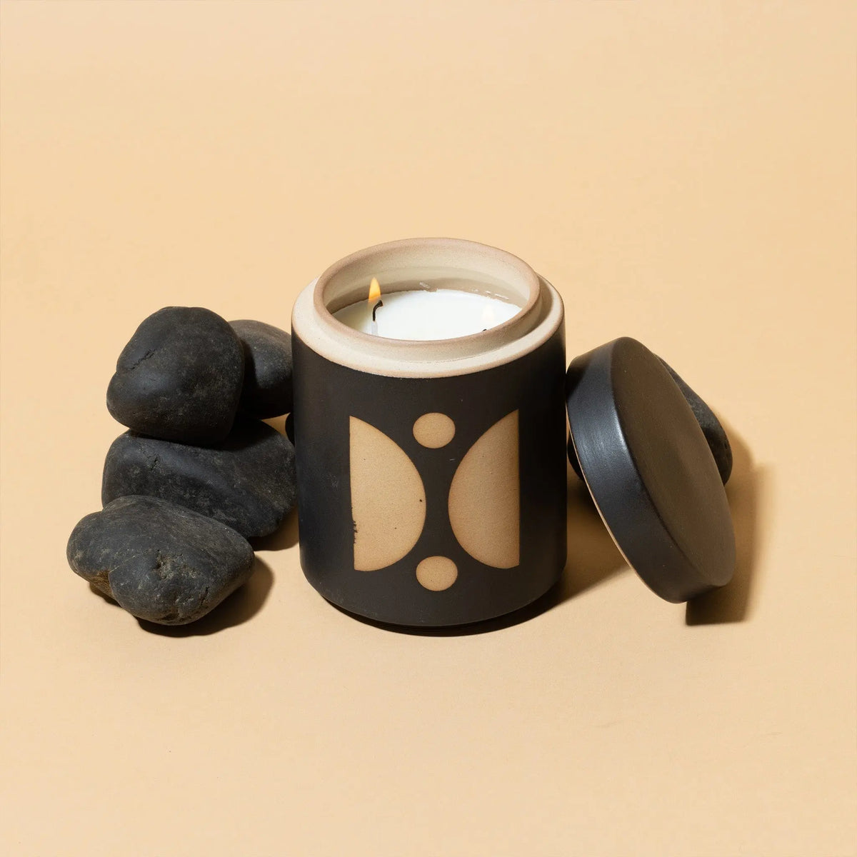 The Palo Santo Suede - Form 12 oz Candle, in a black and beige ceramic jar with its lid off and white candle lit, is displayed beside smooth black stones on a beige background.