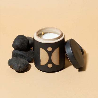The Palo Santo Suede - Form 12 oz Candle, in a black and beige ceramic jar with its lid off and white candle lit, is displayed beside smooth black stones on a beige background.