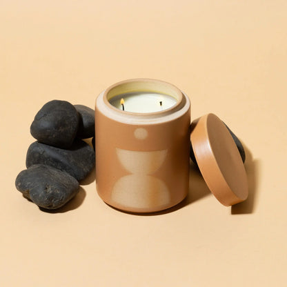The Wild Fig + Vetiver - Form 12 oz Candle, in a geometric ceramic jar, sits beside black stones on a beige surface. Its lid is placed to the right.