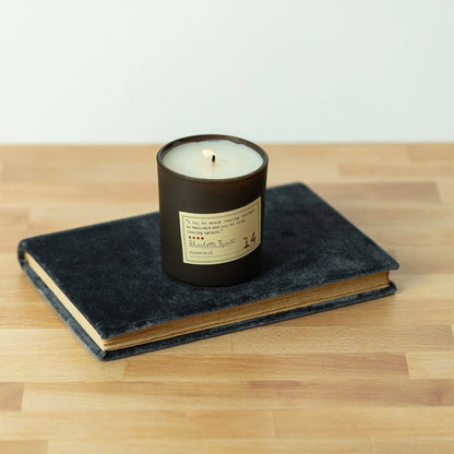 The Charlotte Bronte - Library Candle 6 oz, in a glass jar, rests on a dark hardcover atop wood—a perfect fragrance gift for book lovers from the inviting Library Collection.