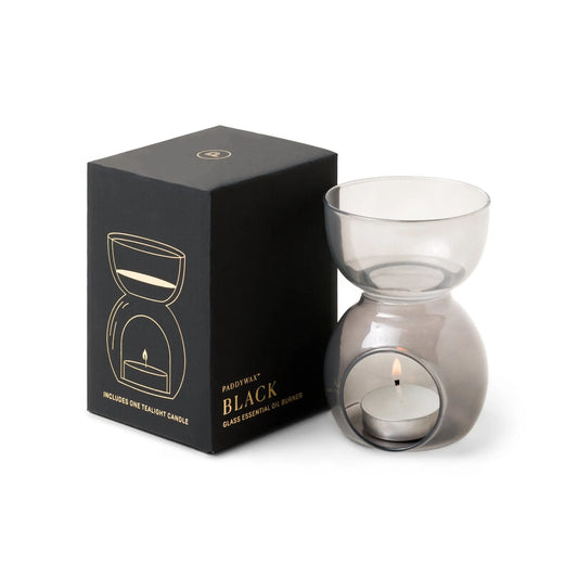 A sleek Black Glass - Essential Oil Burner & Tea Light Candle, illuminated by a glowing tea light inside, sits next to its matching black box adorned with a gold line drawing of the burner and the product name, “Black Glass - Essential Oil Burner & Tea Light Candle.”.
