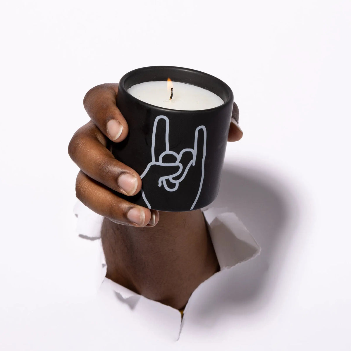 A hand emerges through a torn white surface, holding the Leather + Oakmoss "Rock On" - Impressions 5.75 oz Candle, a matte black candle featuring a rock hand gesture design—perfect for fans of unique and edgy scented decor.