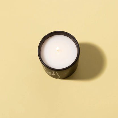 The Leather + Oakmoss "Rock On" Impressions 5.75 oz Candle in a black glass holder is lit and set on a light yellow background, casting a gentle shadow to the right.
