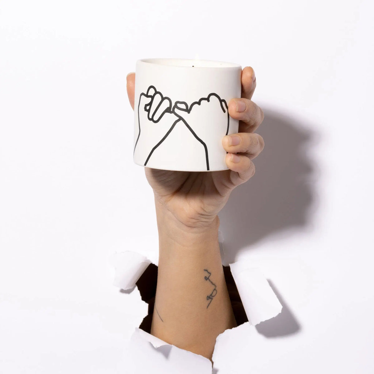 A hand with a small tattoo holds the Wild Fig + Cedar "Pinky Promise" Impressions 5.75 oz Candle, featuring a line drawing of two pinky fingers intertwined on its white surface, emerging from a tear in a white background.
