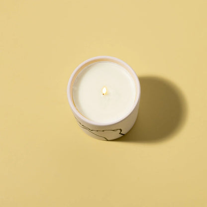 The Wild Fig + Cedar "Pinky Promise" - Impressions 5.75 oz Candle is lit in a round container on a plain beige background, casting a shadow to the right.