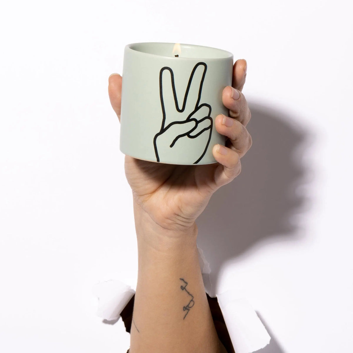 A hand holds the Lavender + Thyme "Peace" Impressions 5.75 oz Candle, featuring a black peace sign outline, emerging through a white background.