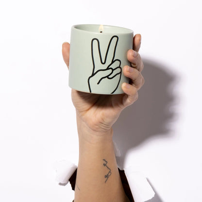 A hand holds the Lavender + Thyme "Peace" Impressions 5.75 oz Candle, featuring a black peace sign outline, emerging through a white background.