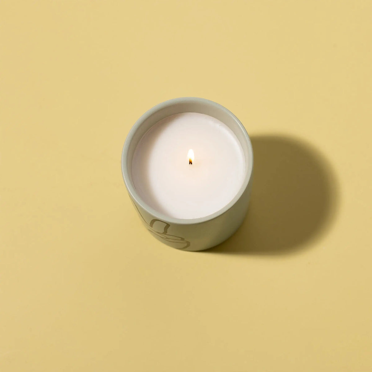 The Lavender + Thyme "Peace" Impressions 5.75 oz Candle glows softly in its ceramic container, its white matte wax burning with a gentle flame and casting a subtle shadow on a plain beige surface.