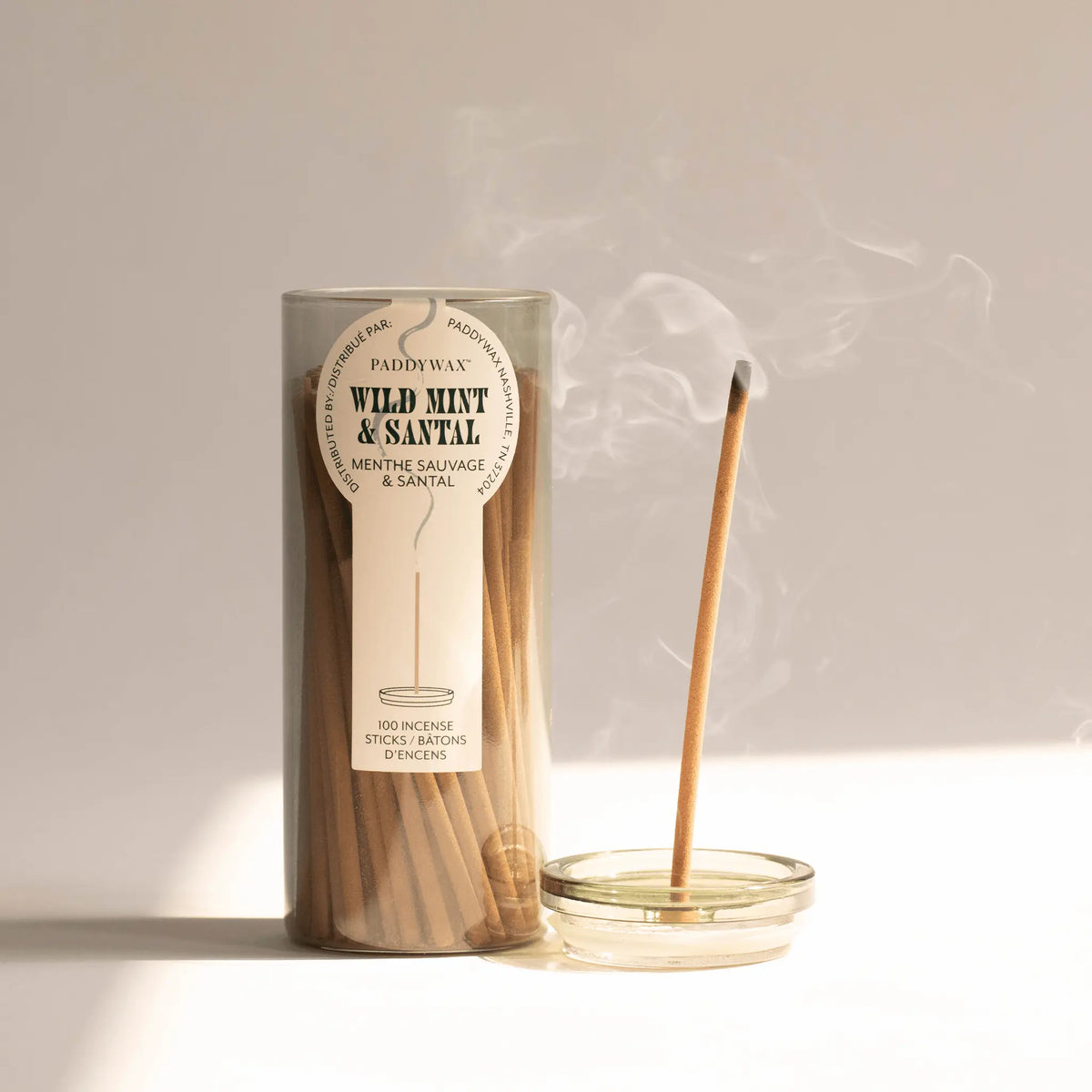 The Wild Mint + Santal - Haze Incense, part of an incense collection, burns in a small glass holder, releasing flameless fragrance and smoke against a neutral background. A matching glass container completes the set.