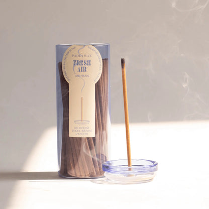 A clear container labeled "Fresh Air - Haze Incense" holds a collection of slender incense sticks. One stick is burning in a small glass incense holder, its flameless fragrant smoke curling in soft sunlight across a light surface.