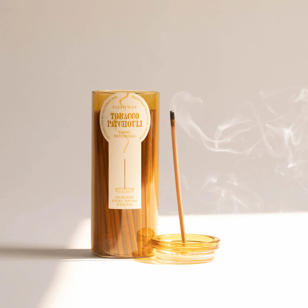 A brown glass jar labeled Tobacco Patchouli - Haze Incense, part of an elegant incense collection, holds fragrant incense sticks. Beside it, a round incense holder supports a lit stick that releases a delicate trail of smokeless fragrance against a light background.