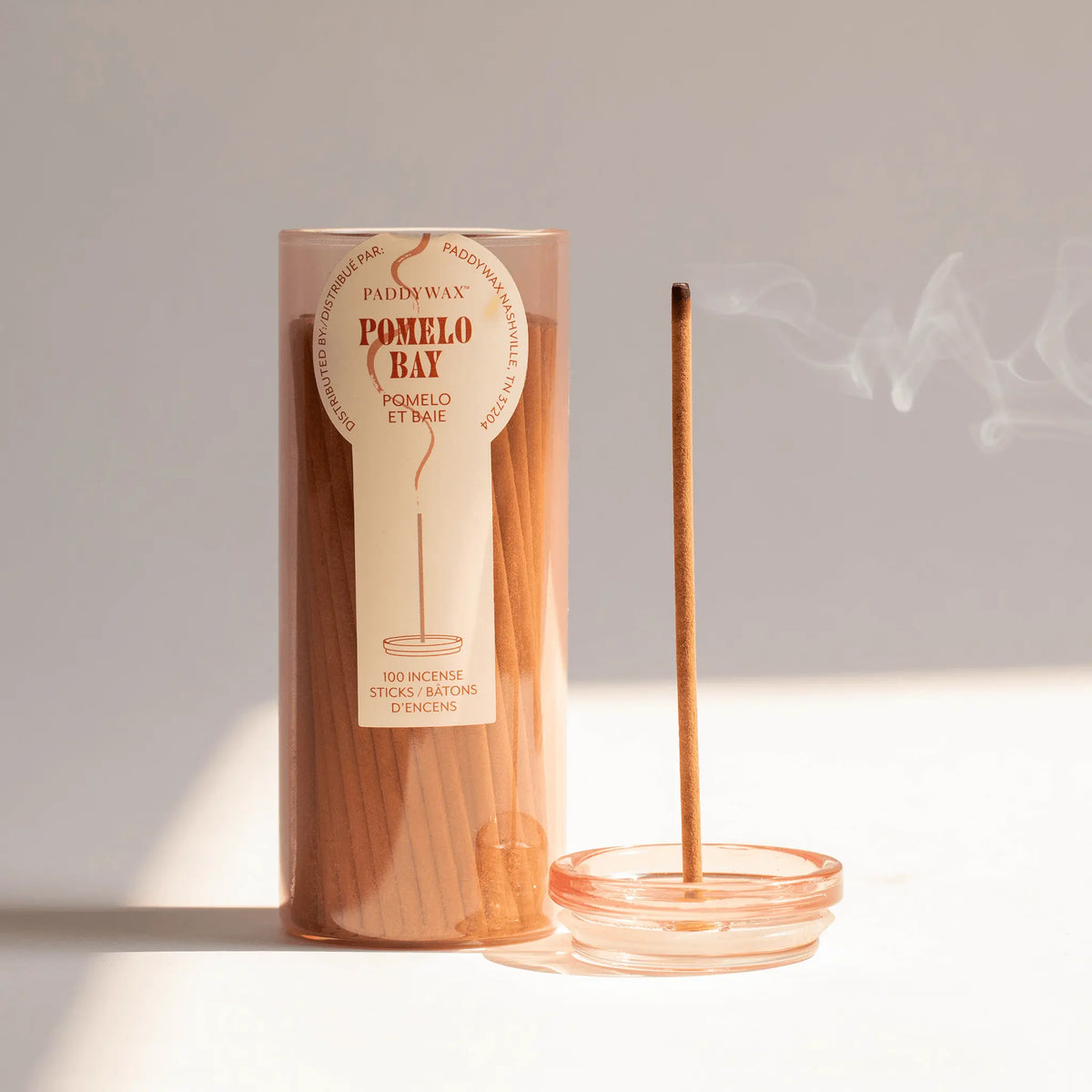 The Pomelo Bay - Haze Incense collection is presented in a cylindrical container. A single incense stick stands lit in a glass holder, with smoke curling gently against a neutral background.