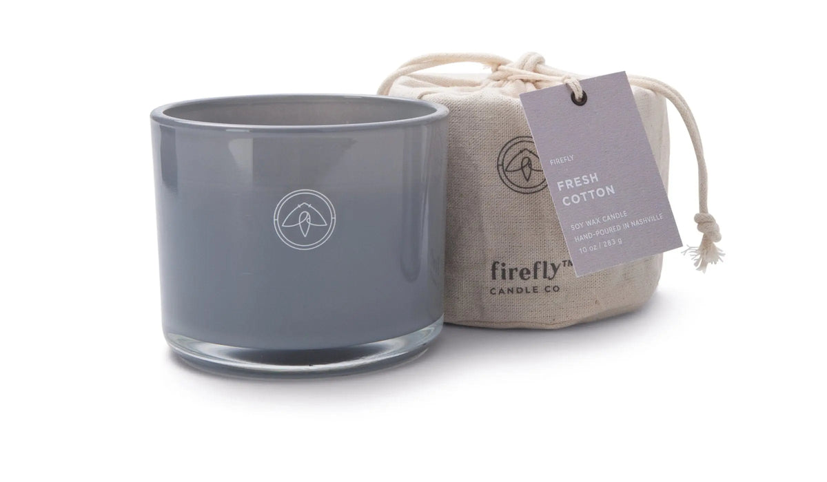 A gray glass “Fresh Cotton - Kindred 10 oz Candle” sits beside a linen bag with a tag reading “Fresh Cotton Soy Wax Candle,” making this candle set perfect for gifting or cozy evenings at home.