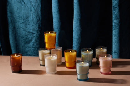Nine lit candles in colorful glass holders are arranged on a beige surface in front of a blue velvet curtain. The glass holders are in white, yellow, orange, pink, and blue tones.