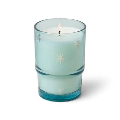 A light blue White Woods & Mint - Noel Candle 5.5 oz burns with a gentle flame in vintage glassware adorned with white star patterns, creating a festive and refreshing ambiance against a white background.