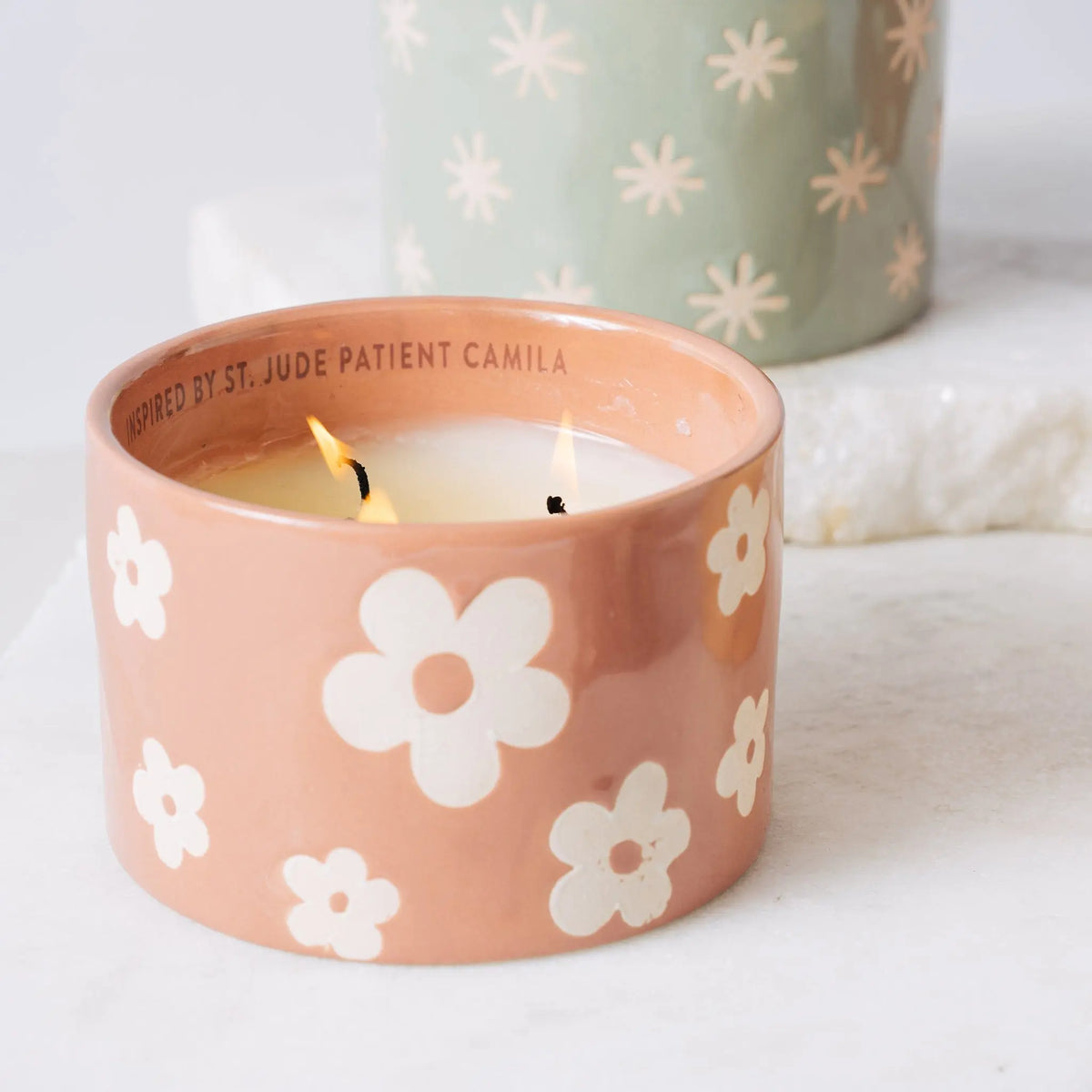 The Sky + Rain - St. Jude Giveback "Hope" 11 oz. Candle, housed in a peach-colored ceramic holder adorned with white flower designs, glows warmly. In the background, a green ceramic holder featuring similar floral patterns is partially visible.
