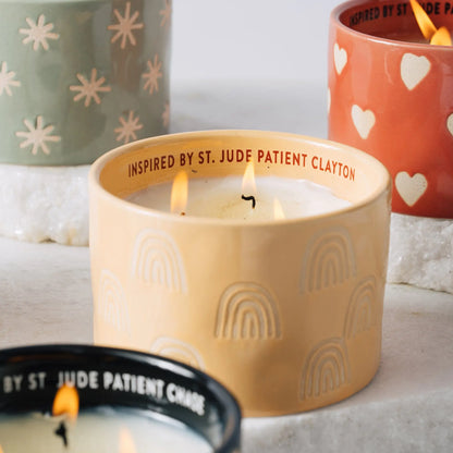 A close-up of four decorative candles in colorful holders. The front yellow Cactus Flower - St. Jude Giveback "Possibilities" 11 oz. Candle is lit, symbolizing hope for childhood cancer patients and inspired by St. Jude patient Clayton.