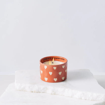 The Rosewater Santal - St. Jude Giveback "Courage" 11 oz. Candle, a small orange ceramic holder with white hearts, rests on a white marble surface against a simple light background, symbolizing support for childhood cancer awareness.