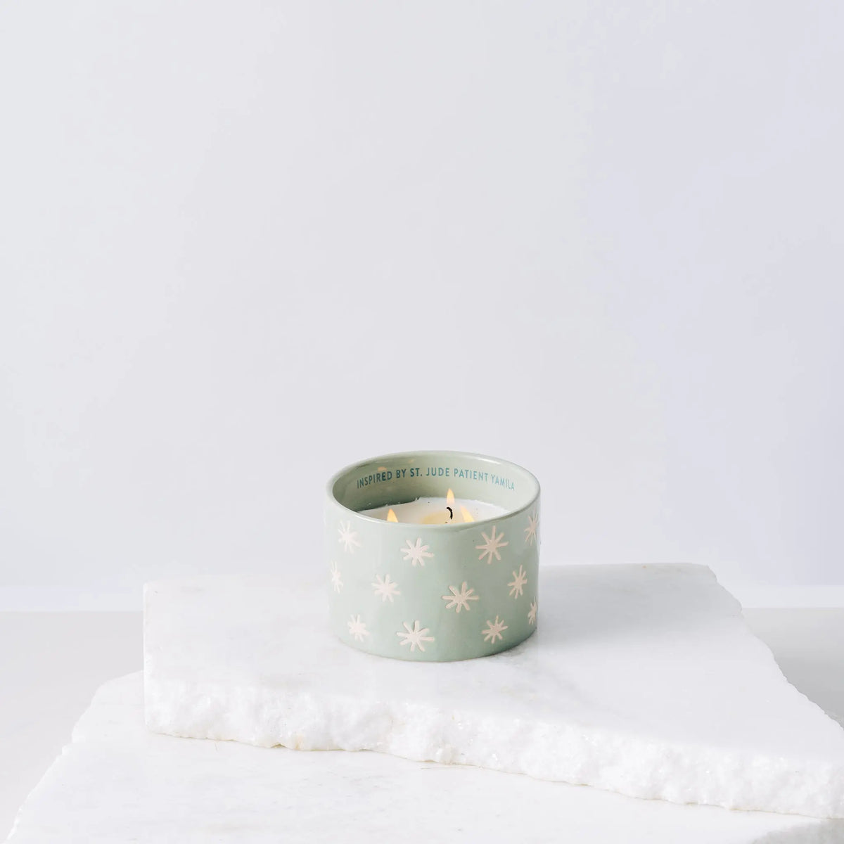 A light green ceramic candle featuring white star patterns, the Mist + Mint - St. Jude Giveback "Inspire" 11 oz. Candle, sits lit on a white marble slab against a plain white background.