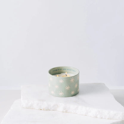 A light green ceramic candle featuring white star patterns, the Mist + Mint - St. Jude Giveback "Inspire" 11 oz. Candle, sits lit on a white marble slab against a plain white background.