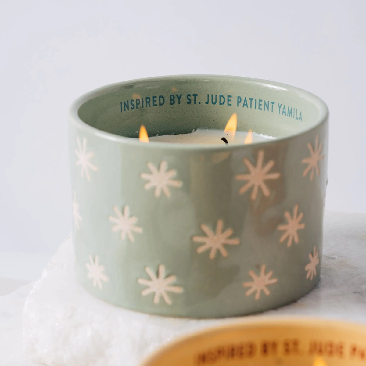 The Mist + Mint - St. Jude Giveback "Inspire" 11 oz. Candle from Paddywax features a light green ceramic holder adorned with white starburst patterns and two glowing wicks. Along the inside rim, the text reads "INSPIRED BY ST. JUDE PATIENT YAMILA." Resting on a white stone surface, this candle blends artistry and inspiration in a beautiful commemorative piece dedicated to St. Jude.