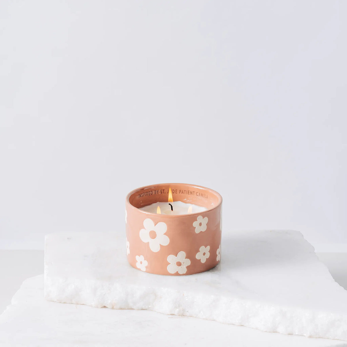 A small, pink ceramic Sky + Rain - St. Jude Giveback "Hope" 11 oz. Candle by Paddywax, adorned with white flower patterns, sits on two stacked white marble slabs. The ceramic candle inside is lit, casting a gentle glow. The background is plain and light.