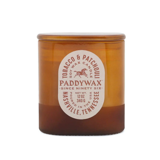 A 12 oz amber glass candle jar from the vintage-inspired Vista collection, the Tobacco + Patchouli - Vista 12 oz Candle features a white label and holds 340g of premium soy wax in a tonal glass vessel.