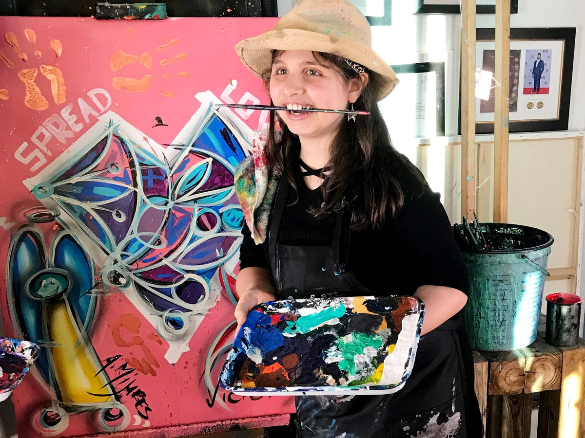 A smiling girl in a hat and black apron holds a paint palette and brush before a colorful "SPREAD LOVE" butterfly painting. Nearby, the Rosewater Santal - St. Jude Giveback "Courage" 11 oz. Candle glows, filling the art-filled room with hope.