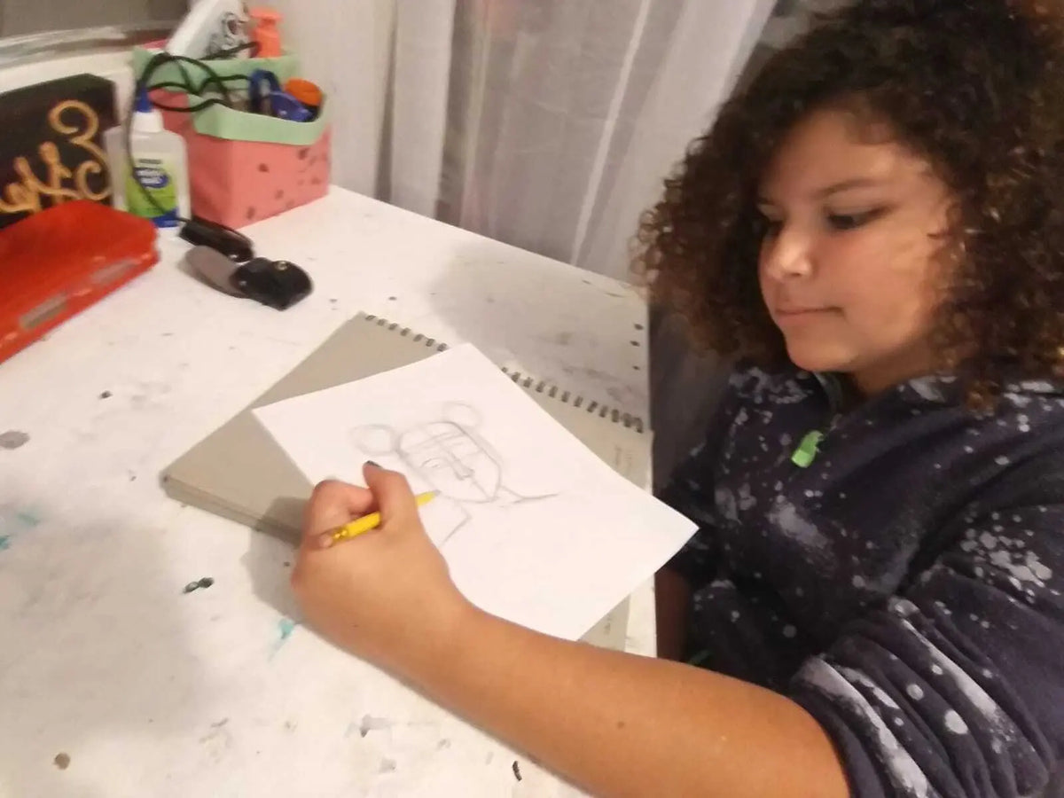 At a cluttered desk, a child with curly hair focuses intently on drawing a bear-eared character on white paper, while the Mist + Mint - St. Jude Giveback "Inspire" 11 oz. Candle flickers softly nearby.
